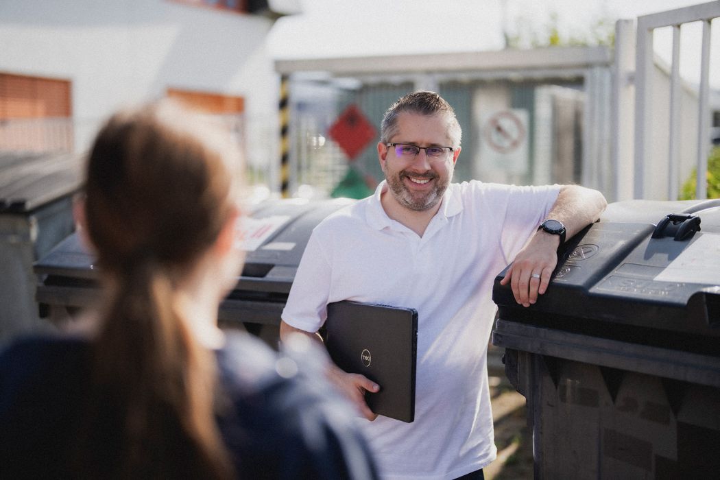 Ein Mann mit Laptop in der Hand spricht mit einer Frau, er lächelt und lehnt sich an einen Müllcontainer.